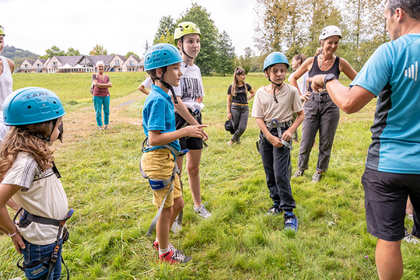 Educateur sportif de dos s'adressant à un groupe d'enfants et d'adultes équipé de casque et baudrier avant de grimper dans un arbre