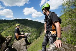 Grimpe :2 hommes l'un face à l'autre sur des rochers à plusieurs mètres d'altitude avec une vue magnifique sur les collines en face